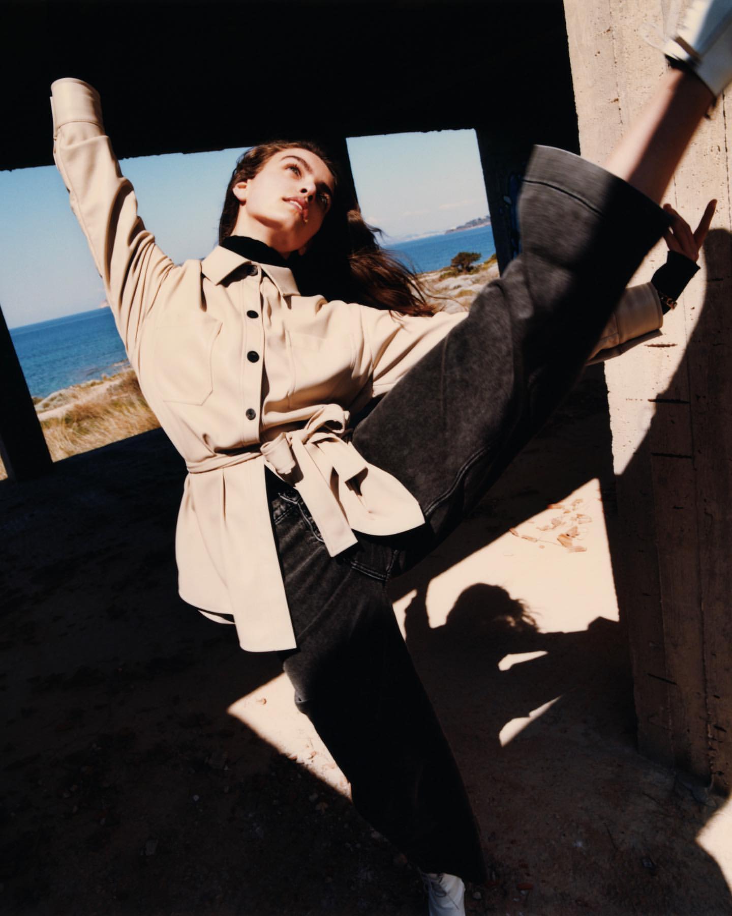 A fashionable adult woman strikes a pose outdoors with a backdrop of the ocean, wearing jeans and a jacket with her arms raised.