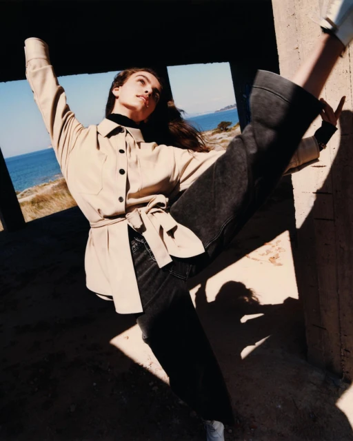 A fashionable adult woman strikes a pose outdoors with a backdrop of the ocean, wearing jeans and a jacket with her arms raised.