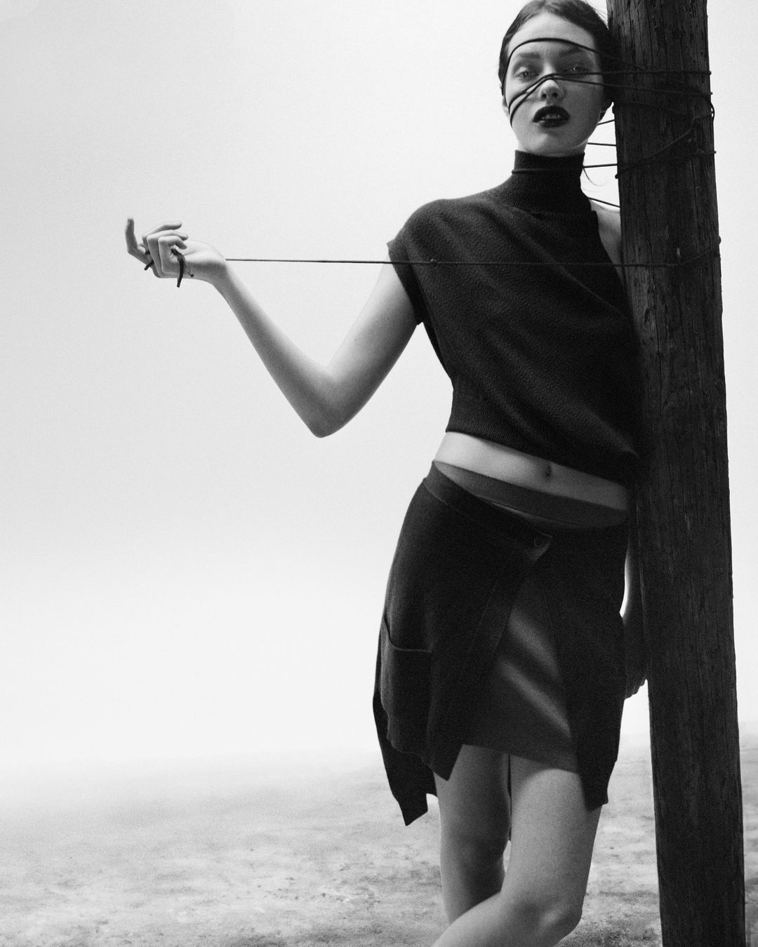 Monochrome studio shot of woman in dark apparel leaning on wooden post, with string wrapped around her face and holding the end.