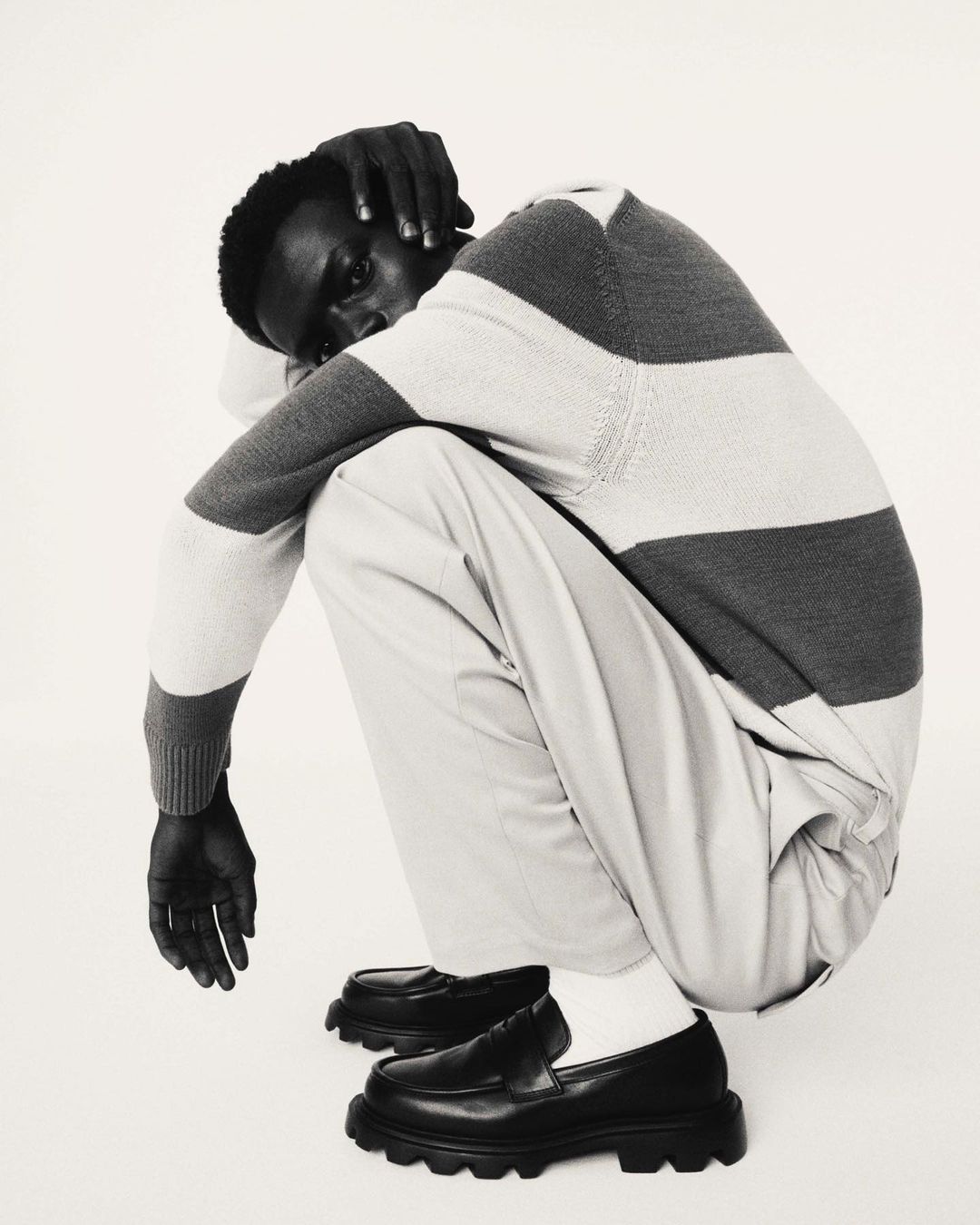 Monochrome portrait of an African-American male model in a striped sweater and loafers squatting against a white studio backdrop.