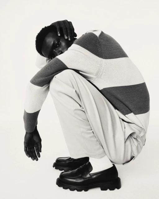 Monochrome portrait of an African-American male model in a striped sweater and loafers squatting against a white studio backdrop.