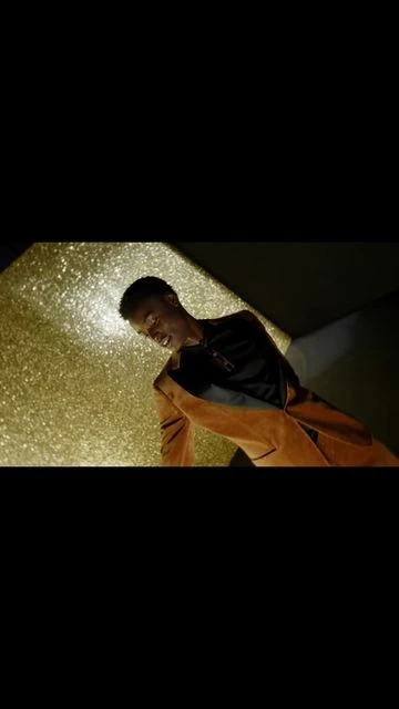 Person in orange and black jacket with gold glittery background looking down in a dimly lit room, close-up shot.