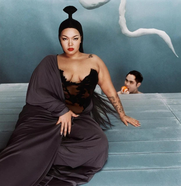 Asian woman in black bustier and gray robe sits in front of a light blue background with a man eating fruit in the background.