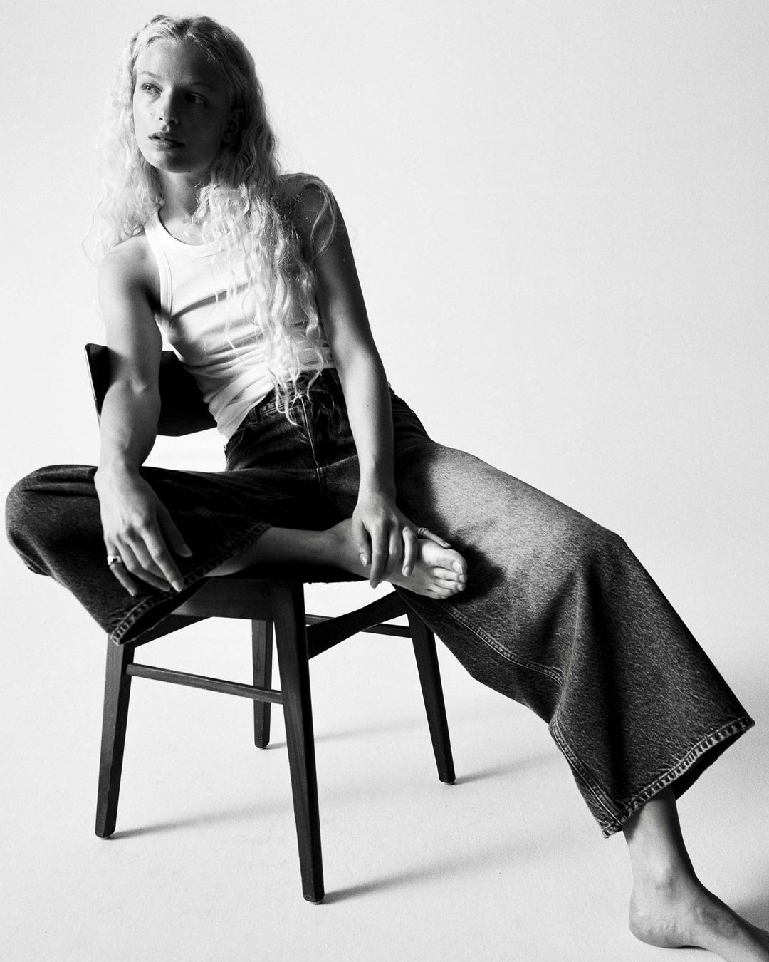 Model with long curly hair wearing a white tank top and wide-leg jeans seated on a dark chair against a white background.