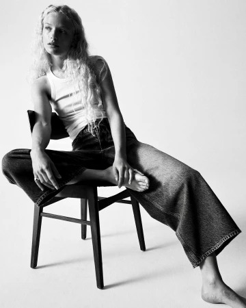 Model with long curly hair wearing a white tank top and wide-leg jeans seated on a dark chair against a white background.