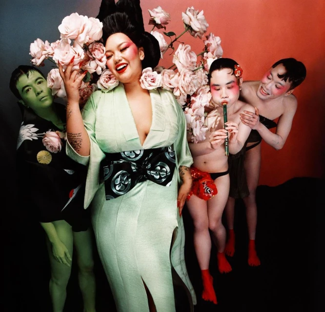 Group of people in theatrical makeup and costumes, posing with pale pink roses in a studio with dark background and bright makeup.