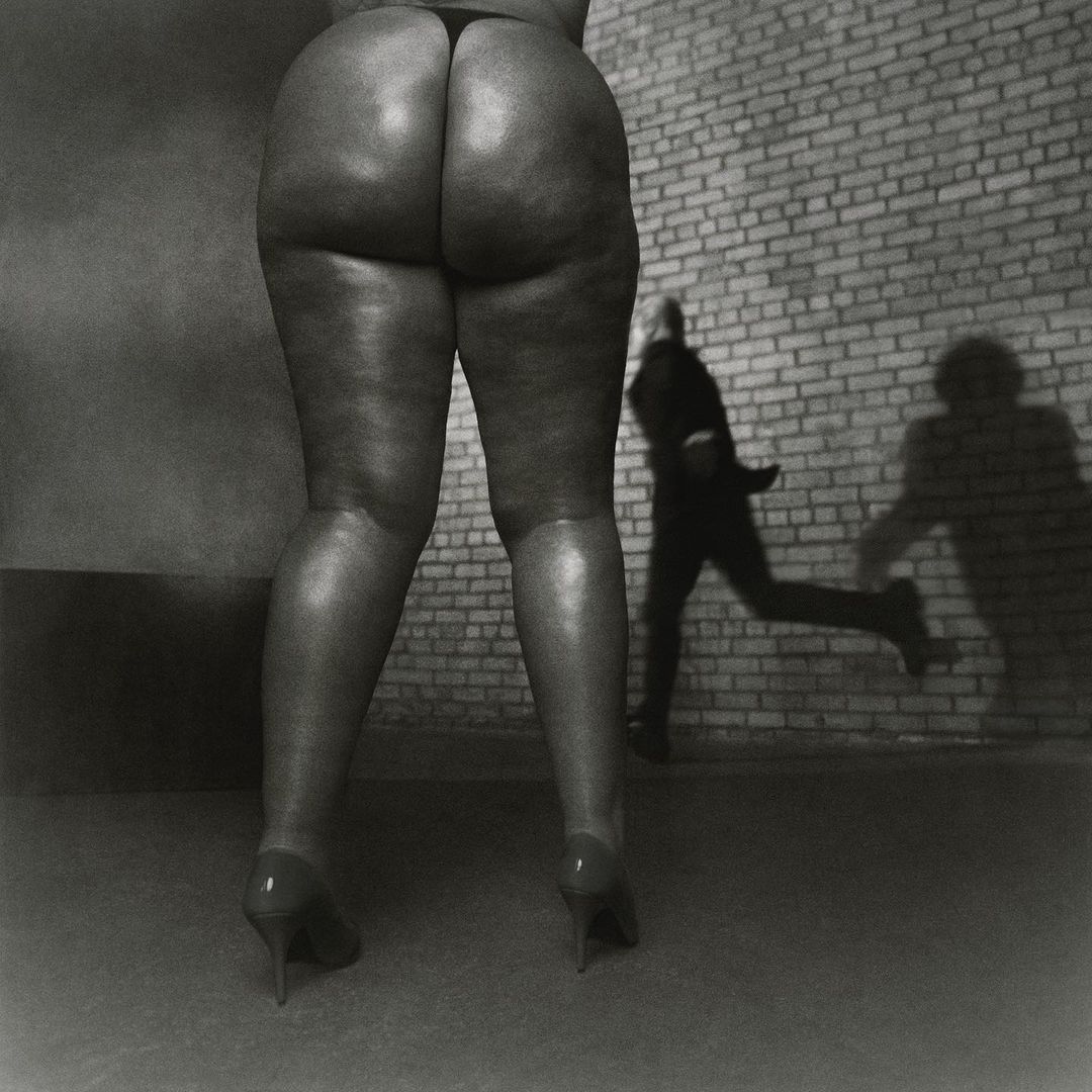 Back view of a person's legs and buttocks in heels against a brick wall with a blurred man running in the background.