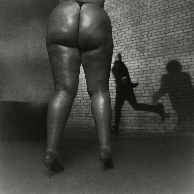 Back view of a person's legs and buttocks in heels against a brick wall with a blurred man running in the background.