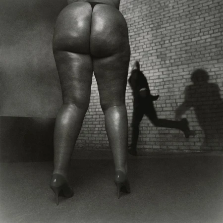 Back view of a person's legs and buttocks in heels against a brick wall with a blurred man running in the background.