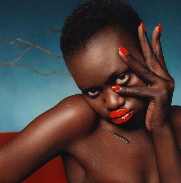 Close-up portrait of a dark-skinned woman with short hair, red lipstick and nails, posing with her hand near her face, on a blue background.