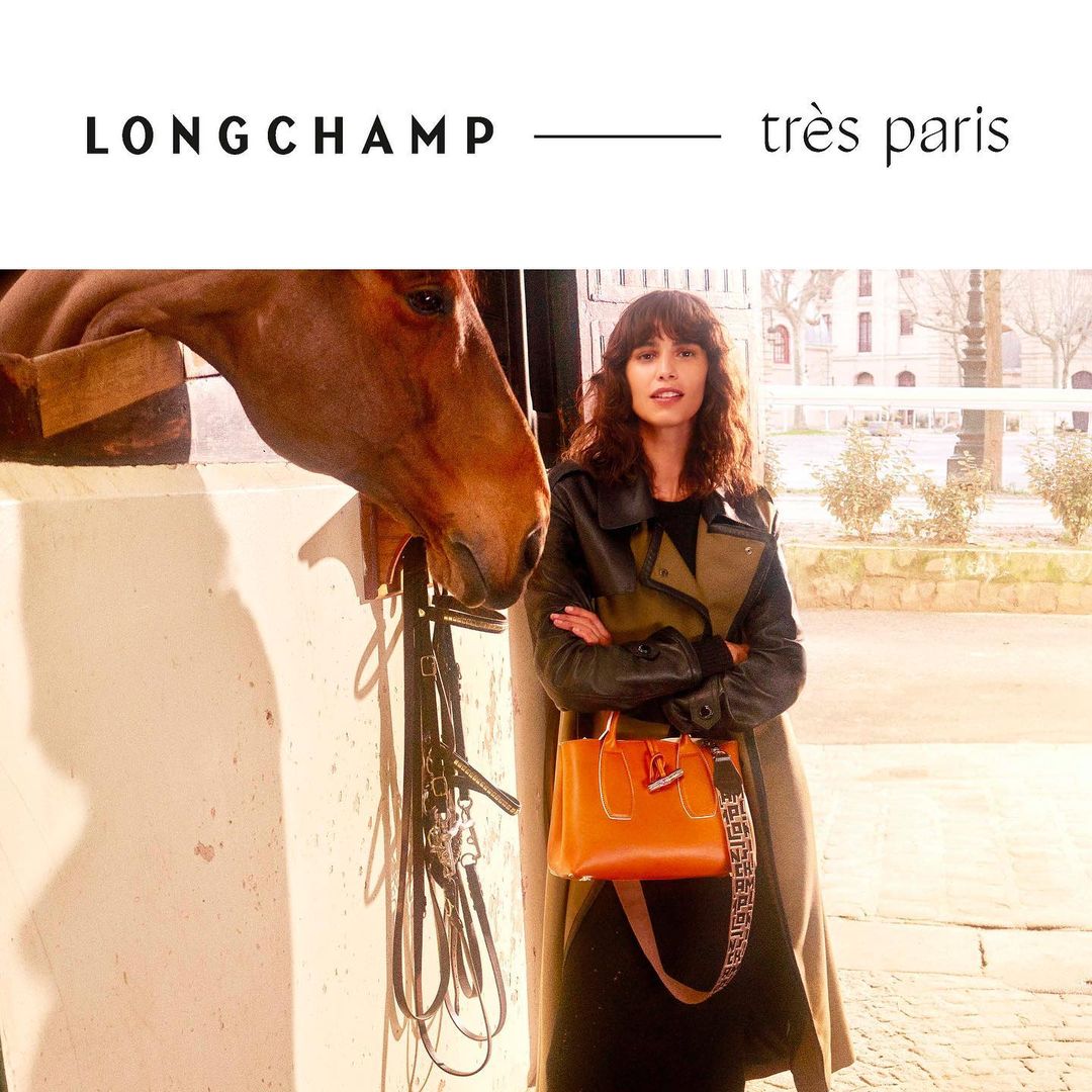 A woman poses with a horse near a building in an outdoor fashion shot. The woman wears a coat and holds a Longchamp purse.
