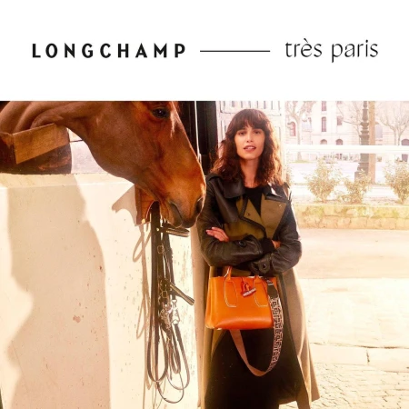 A woman poses with a horse near a building in an outdoor fashion shot. The woman wears a coat and holds a Longchamp purse.