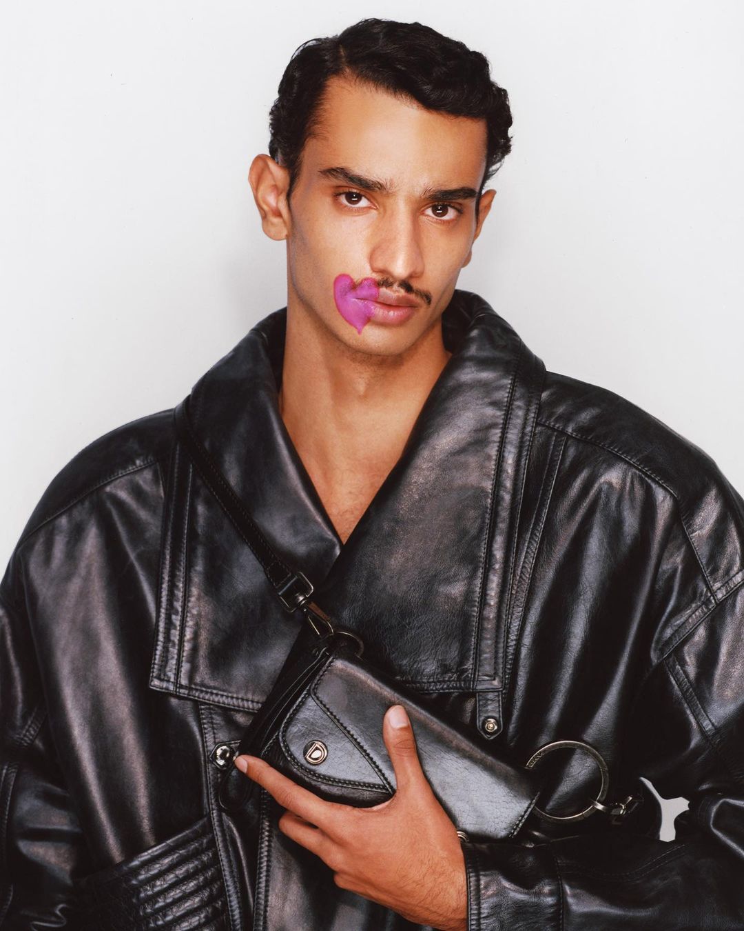 Stylish man in a black leather jacket holds a clutch bag, accented with a pink heart graphic near his lip, posing against a white backdrop.
