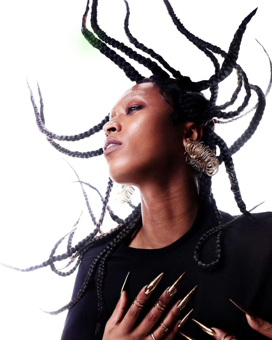 Close-up of a Black woman with braided hair, wearing gold jewelry and long gold fingernails, against a white background. Beauty shot.