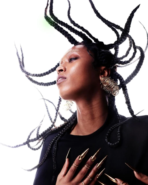 Close-up of a Black woman with braided hair, wearing gold jewelry and long gold fingernails, against a white background. Beauty shot.