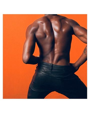 A muscular adult male model with dark skin and shiny leather pants poses against a solid orange backdrop, showcasing physique.