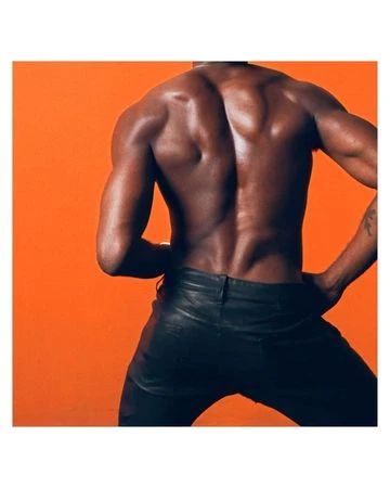 A muscular adult male model with dark skin and shiny leather pants poses against a solid orange backdrop, showcasing physique.