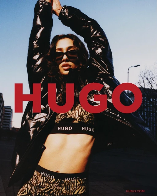 Fashion model poses in animal-print set and black jacket for a HUGO brand ad campaign against an urban backdrop and blue sky.