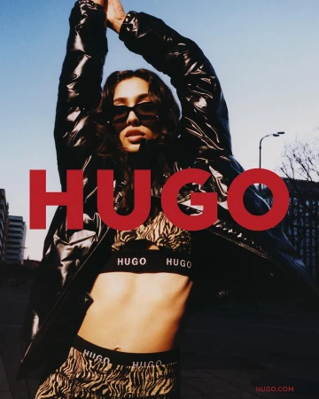 Fashion model poses in animal-print set and black jacket for a HUGO brand ad campaign against an urban backdrop and blue sky.