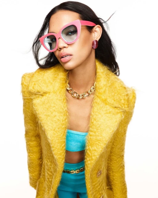 Woman in pink sunglasses and a yellow coat poses against a white background, showcasing fashion and style in a studio shot.