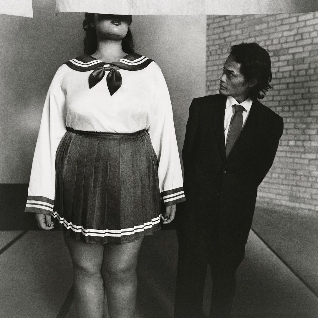 Black and white shot of a person in school uniform with obscured face, and a man in a suit glancing at the person, indoor setting.