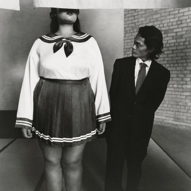 Black and white shot of a person in school uniform with obscured face, and a man in a suit glancing at the person, indoor setting.