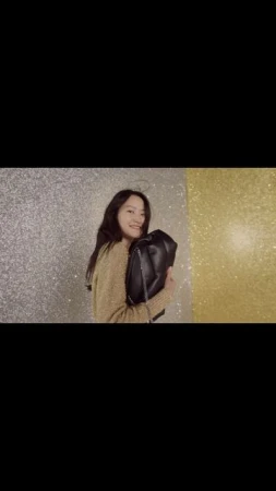 Smiling woman holds a black leather handbag against a shimmering gold and silver background in a close-up fashion shot.