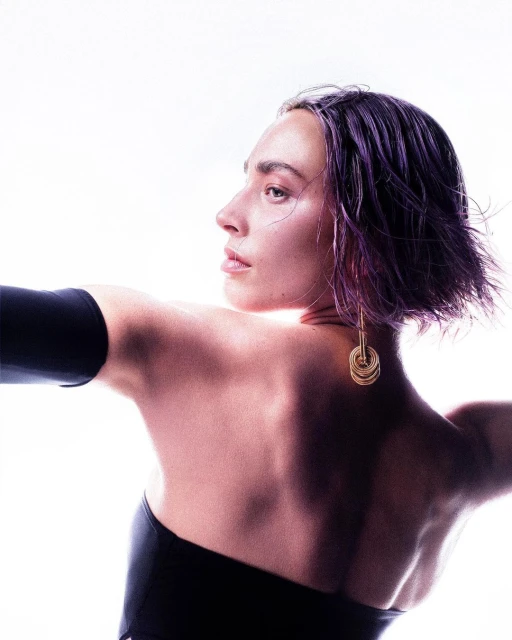Elegant portrait of a woman with purple hair and a black top, looking over her shoulder with a bright, high-key background and golden earrings.