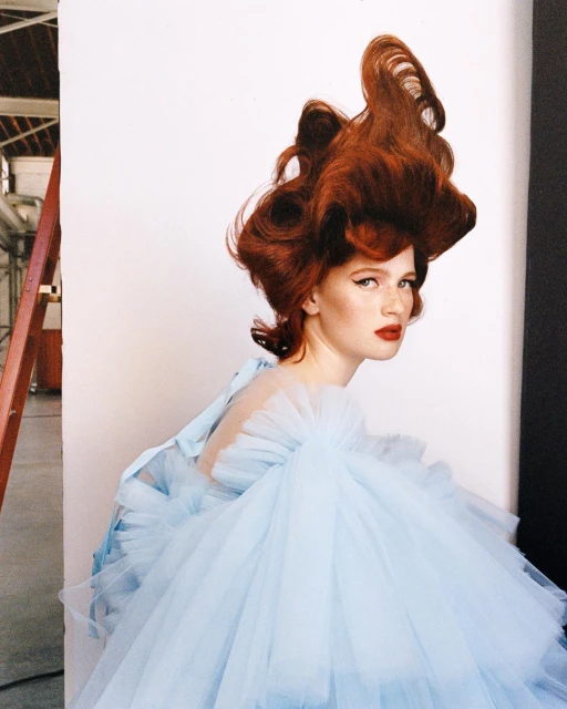 Elegant woman with red hair in a light blue dress posing against a backdrop, showcasing high fashion and sophisticated style indoors.