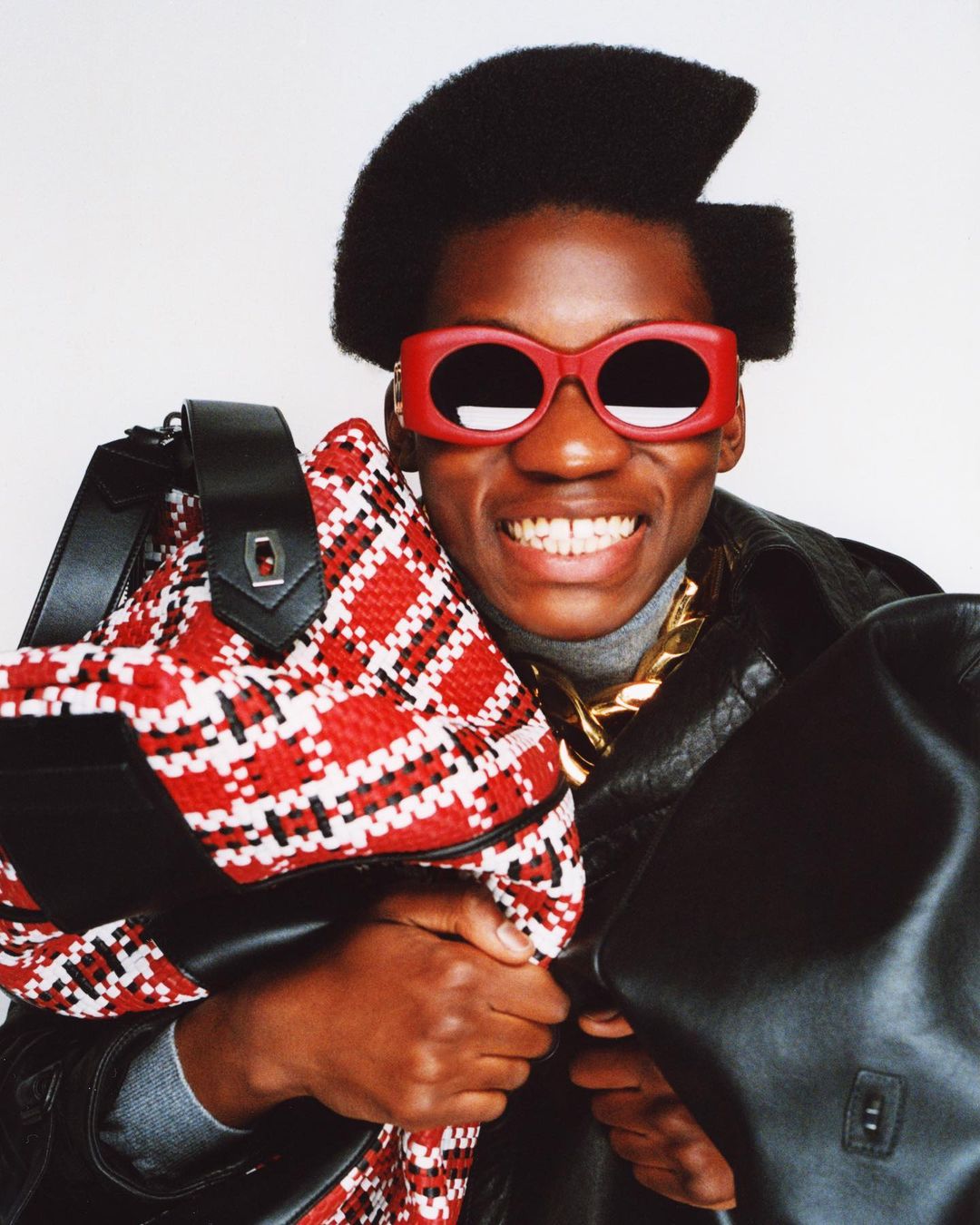 A smiling Black adult male with an afro wearing red sunglasses and a leather jacket is holding two patterned handbags in a bright indoor space.
