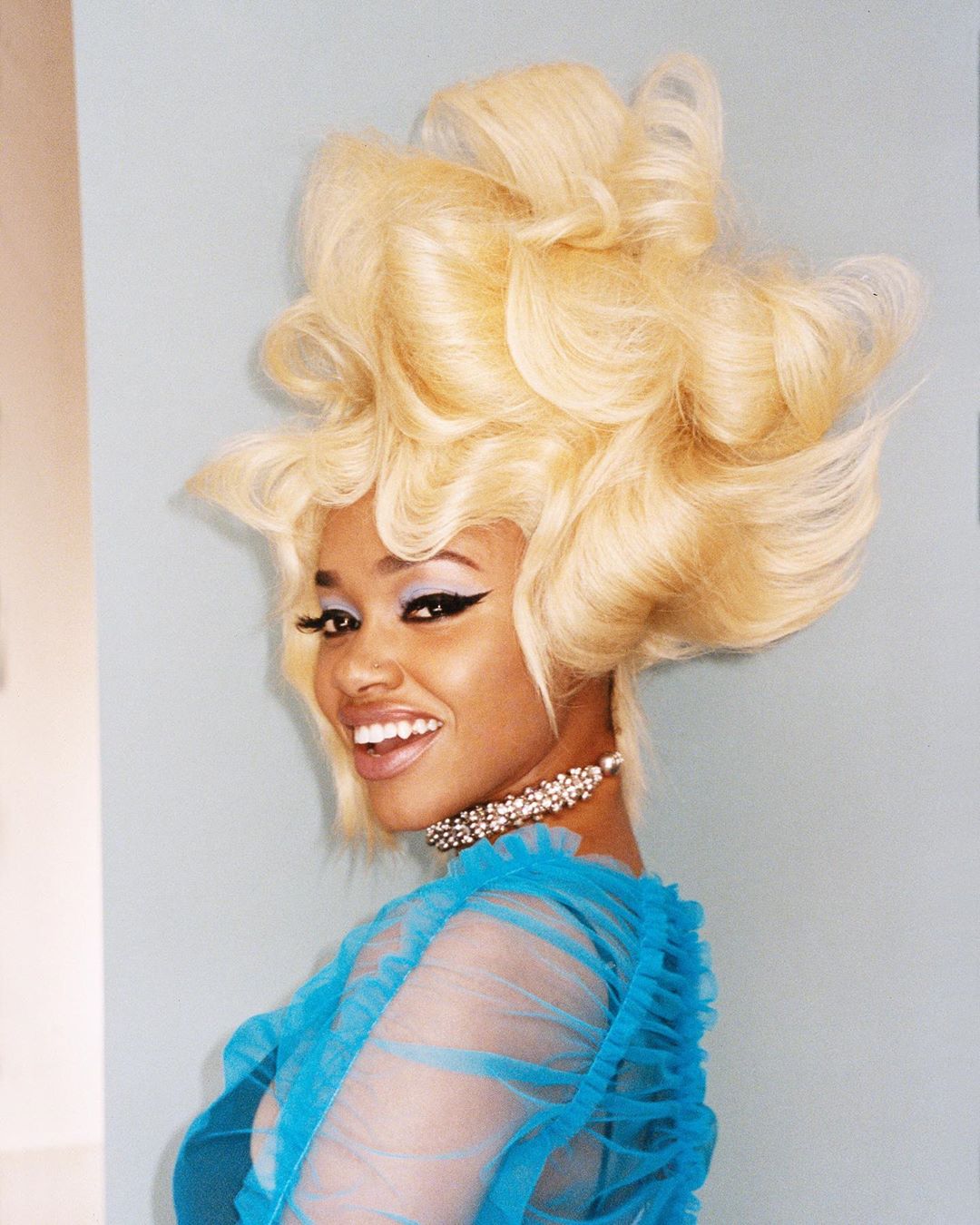 Stunning studio portrait of a smiling young Black woman with elaborate blonde hair, heavy winged eyeliner, and a light blue gown.