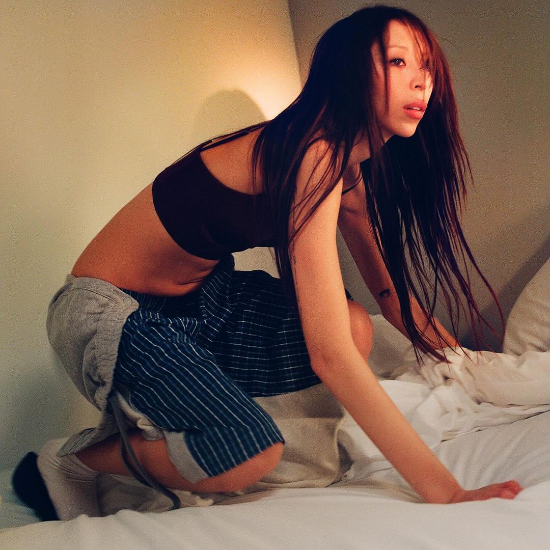 Woman crouches on a bed indoors, wearing a sports bra and shorts. She has long dark hair, fair skin, and faces toward the right.