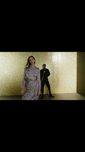 A woman in a patterned dress and a man in a black jumpsuit pose against a gold backdrop, studio lit and fashionable.