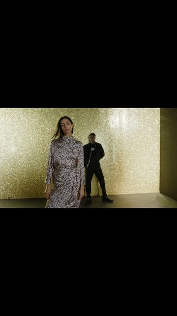 A woman in a patterned dress and a man in a black jumpsuit pose against a gold backdrop, studio lit and fashionable.