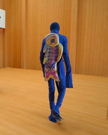 A person in a blue outfit walks away, featuring a large, colorful fish-shaped backpack, against a backdrop of wood paneling.