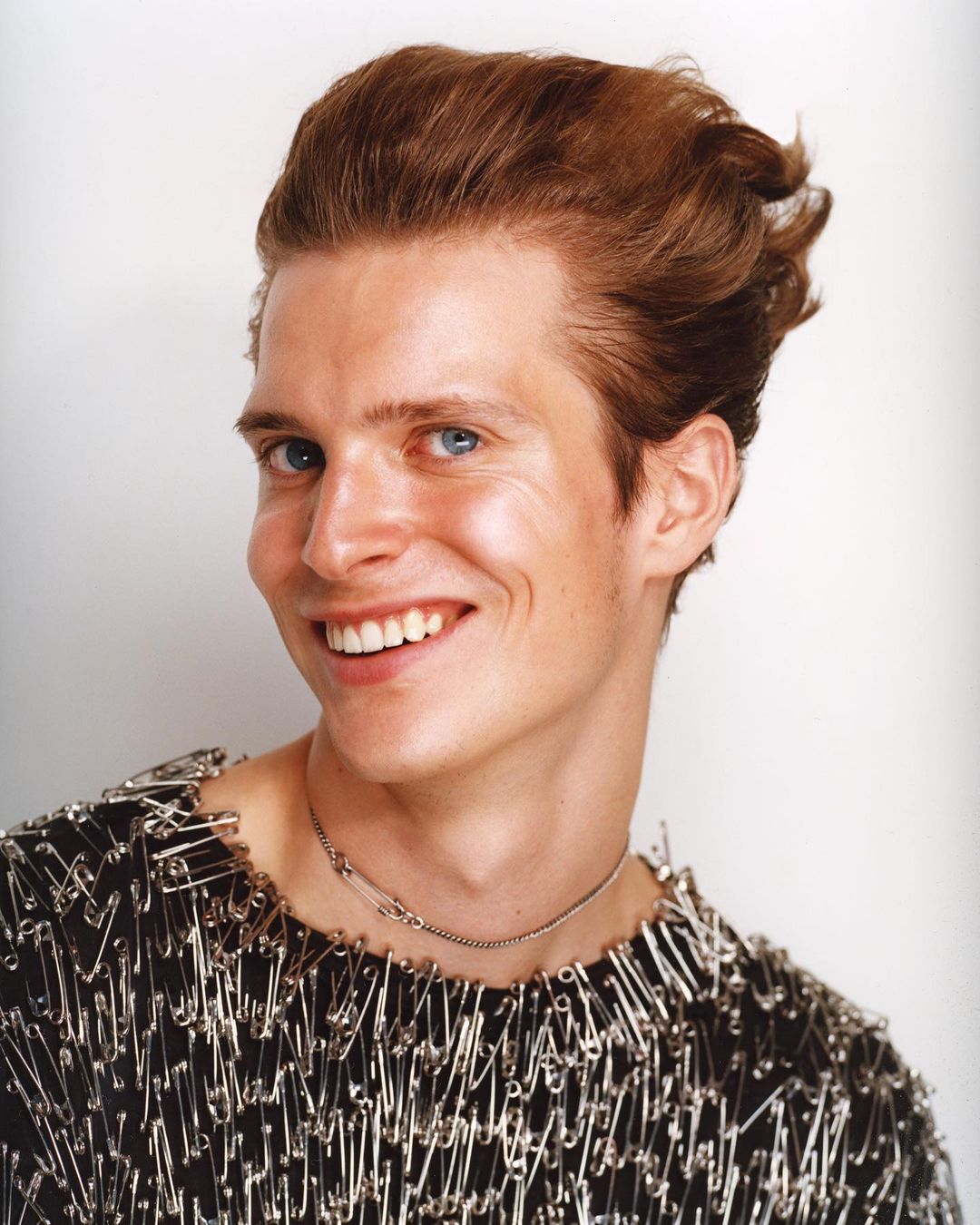 Portrait of a smiling adult male with red hair, wearing a safety pin-covered shirt and necklace, against a white background.