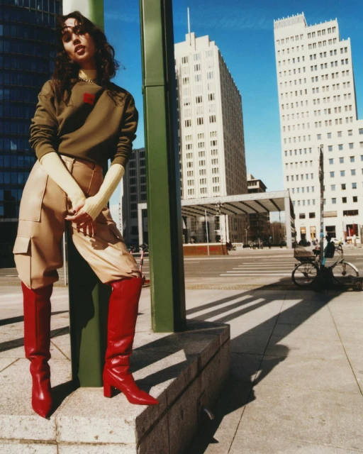 A young woman poses on stone steps in an urban setting, wearing red heeled boots, beige pants, a green sweatshirt, and sunglasses.