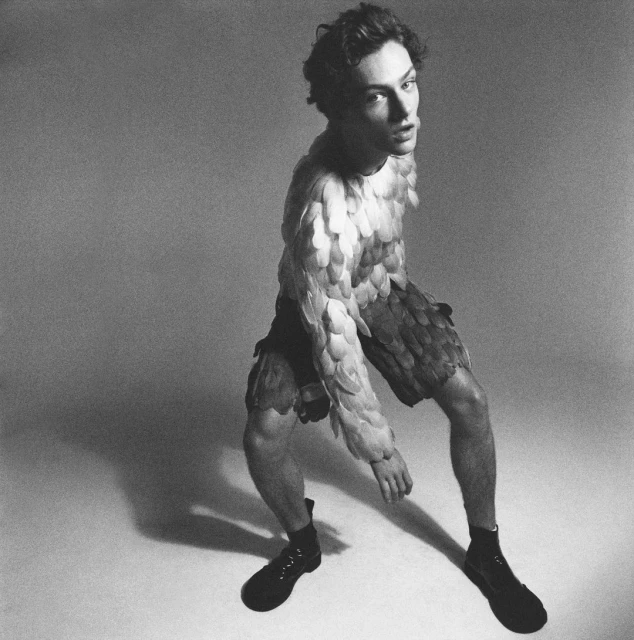 Male fashion model in feathered suit posing in studio. Man wearing feathers, dark boots and looking toward the camera. Black and white photo.