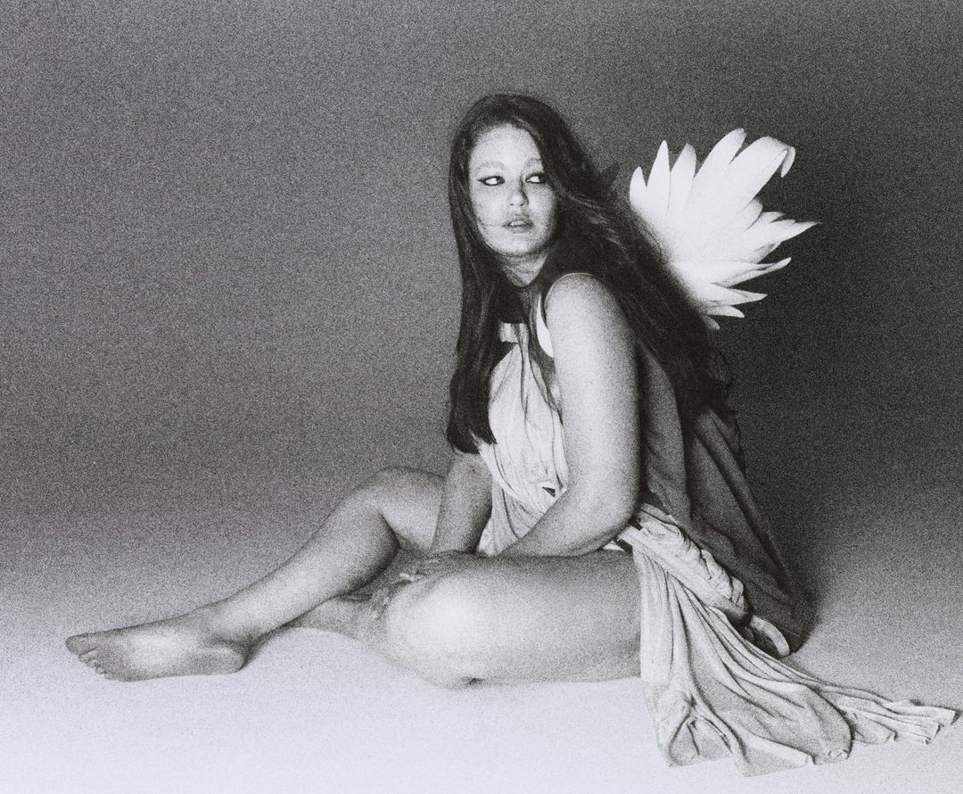 Young woman with long brown hair sits draped in fabric, with feather wings attached to her back in a studio setting. Black and white tones.