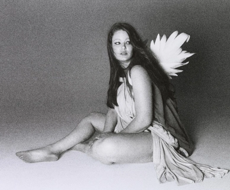 Young woman with long brown hair sits draped in fabric, with feather wings attached to her back in a studio setting. Black and white tones.