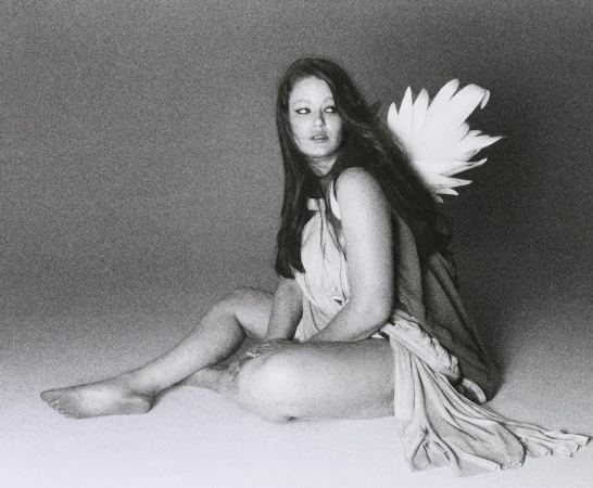 Young woman with long brown hair sits draped in fabric, with feather wings attached to her back in a studio setting. Black and white tones.