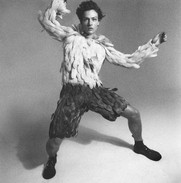 Full shot of adult male dancer in studio setting, wearing unique feathered costume, posed with arms raised and legs in ballet stance.