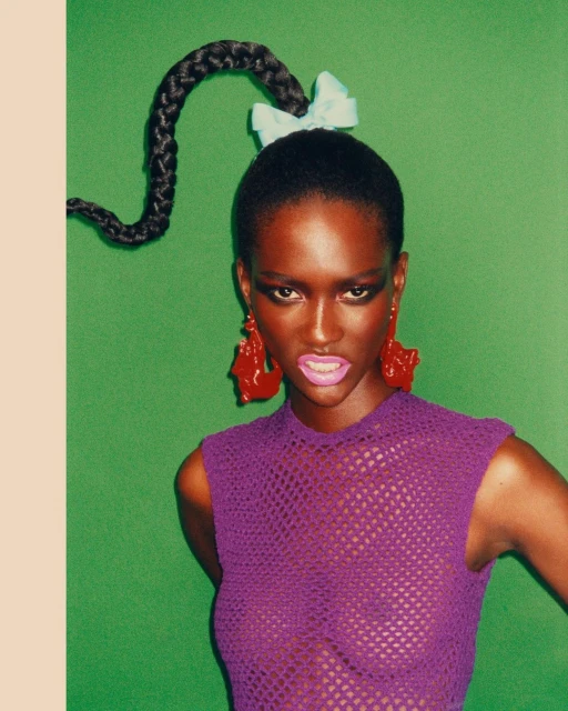 Woman with braided ponytail, red earrings, and a purple mesh top poses against a vibrant green backdrop in a studio setting.