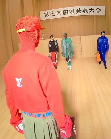 Fashion runway show featuring diverse models in patterned ensembles. A person wearing a red balaclava and sweater faces the stage.