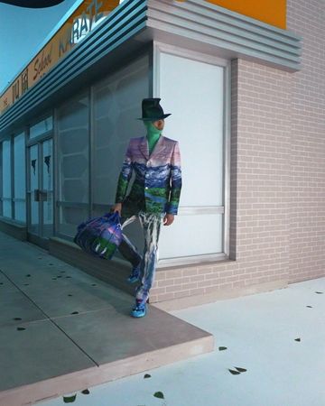 A person stands outside a building in a blazer and pants with a mountain print. They carry a purple duffle bag.