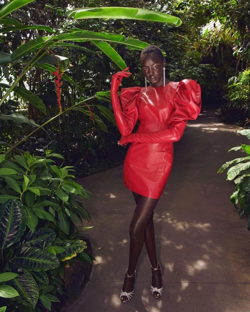 A solo adult female model stands in a lush, tropical green garden wearing a short red dress, matching red gloves, and strappy silver high heels.