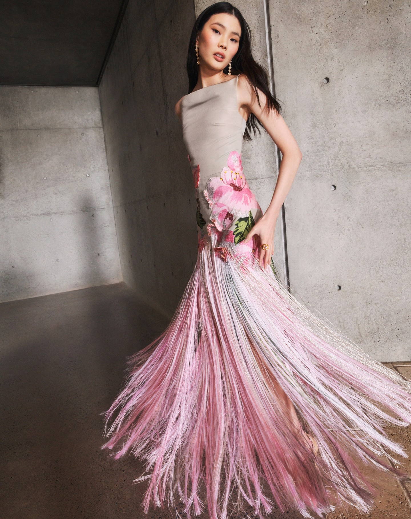 Elegant woman in a sleeveless gown with a floral design poses in front of a concrete wall, showcasing fashion and style with grace and poise.