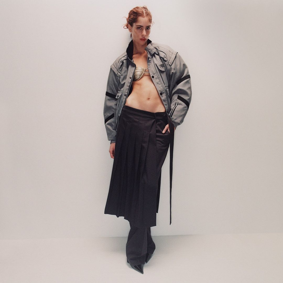 Model with reddish hair wearing an open grey jacket and pleated dark skirt against a plain white background