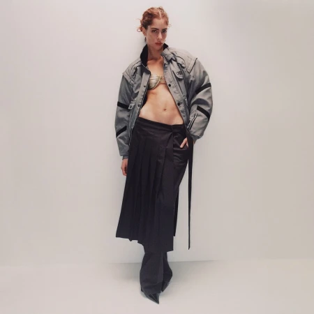 Model with reddish hair wearing an open grey jacket and pleated dark skirt against a plain white background