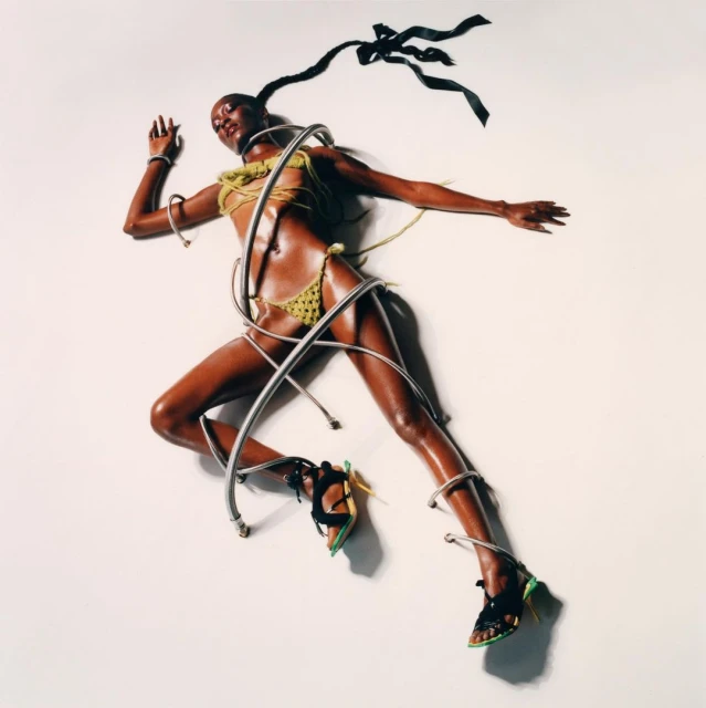 A woman in a green bikini and heels poses on a white background, silver tubing wrapped around her, highlighting fashion and studio photography.
